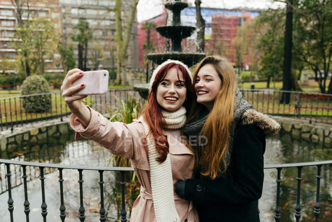 Dos jóvenes amigas guapas paradas en la fuente en el parque y tomando selfie . - foto de stock