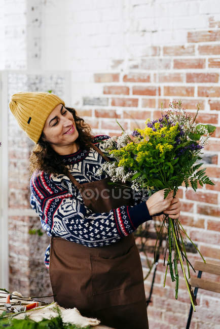 Retrato de florista em camisola de malha e chapéu olhando para buquê nas mãos — Fotografia de Stock