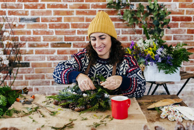 Портрет улыбающейся женщины, сидящей за столом с красной чашкой и делающей рождественский венок — стоковое фото
