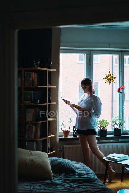 Side view of woman in shirt reading book by window at home — Stock Photo