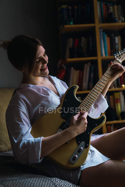 Усміхнена молода жінка сидить і грає на електрогітарі вдома . — стокове фото