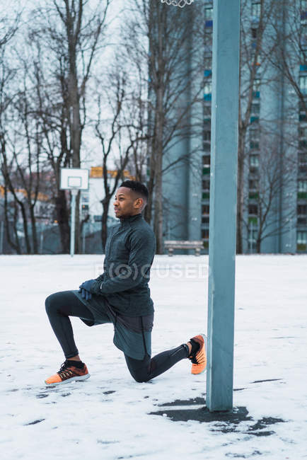 Вид сбоку на черного спортсмена в теплой одежде, растянувшегося на детской площадке зимой — стоковое фото
