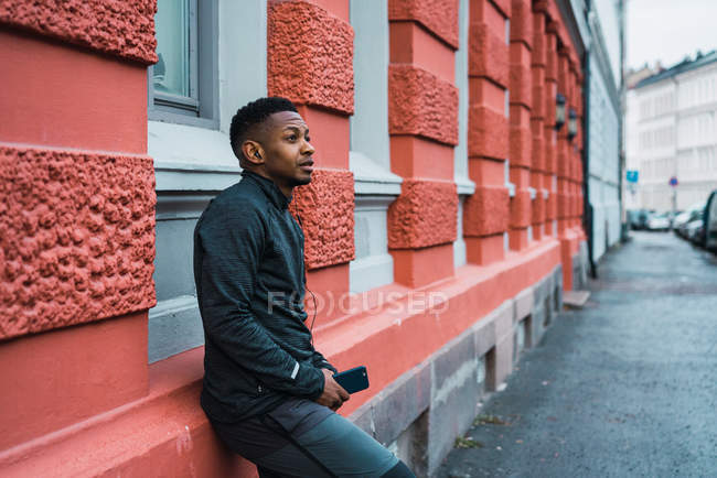 Side view of athletic man in warm sportswear leaning on wall with smartphone in hands and looking away — Stock Photo