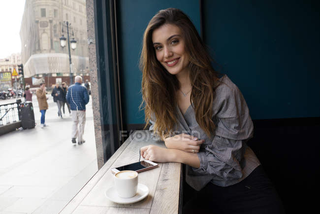 Retrato de una joven morena sentada en la mesa del café con café y smartphone y mirando a la cámara - foto de stock
