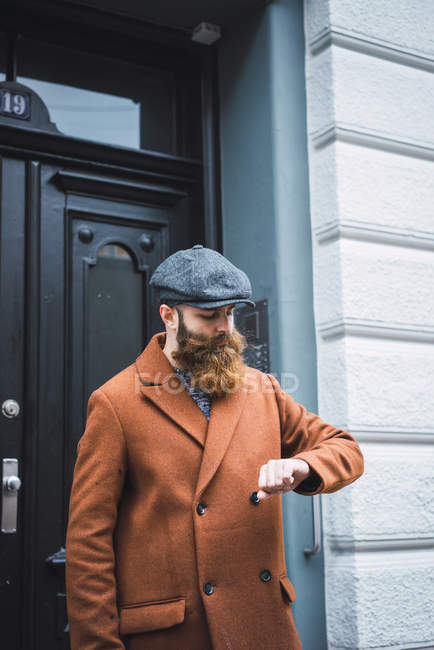 Retrato del hombre barbudo reflexivo mirando el reloj en la mano cerca de la puerta de entrada - foto de stock