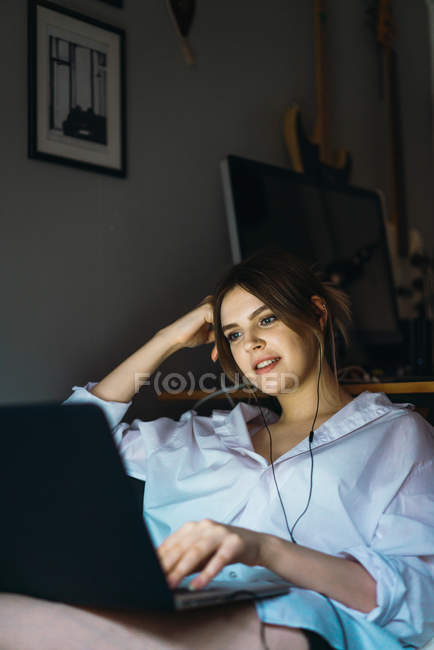 Portrait of cheerful woman in earphones looking at laptop while lying on sofa — Stock Photo