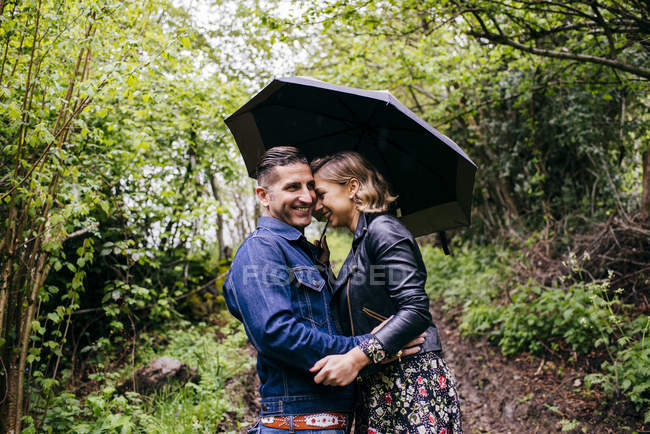 Feliz pareja abrazando mientras de pie bajo paraguas en verde bosque . - foto de stock