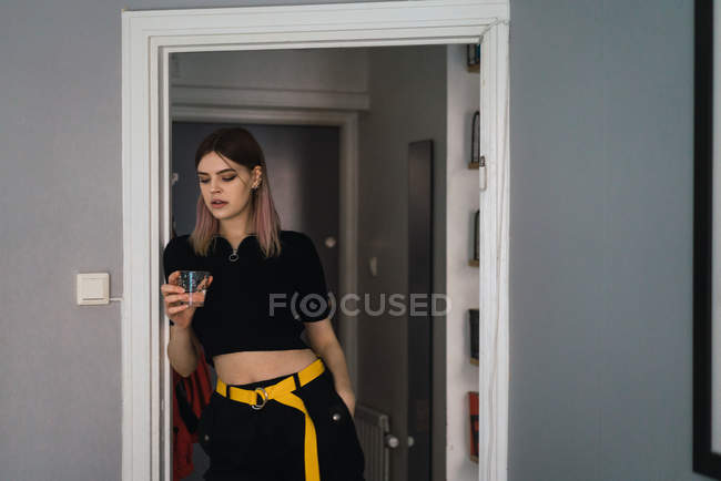 Портрет молодої жінки, що стоїть у дверях зі склянкою води і дивиться вниз — стокове фото