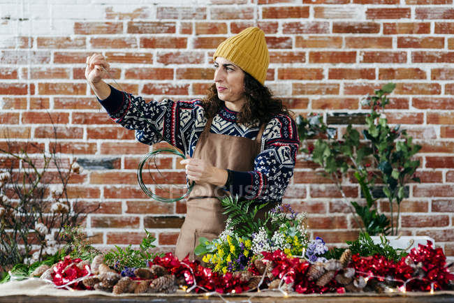 Mujer vista lateral en sombrero y delantal sobre jersey de pie en la mesa con flores y tomando cinta para atar ramo . - foto de stock