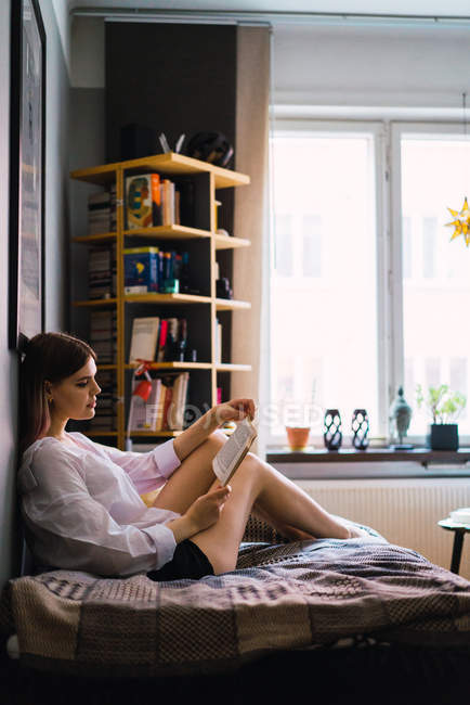 Vista lateral da mulher sentada na cama em casa e livro de leitura — Fotografia de Stock