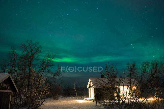 Exterior view of small wooden building in winter forest on background of northern lights in sky — Stock Photo
