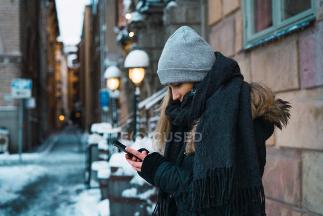 Side view of pretty woman in casual warm clothes using smartphone on city street. — Stock Photo