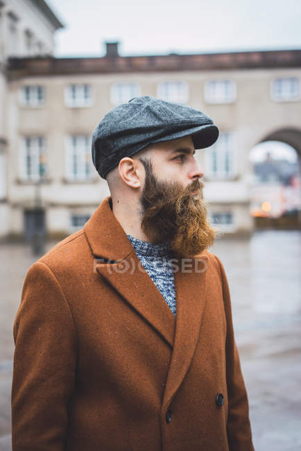 Vista laterale dell'uomo barbuto in berretto e cappotto in posa nella piazza della città — Foto stock