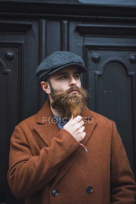 Portrait of thoughtful bearded man in stylish cap and coat palming beard and looking away — Stock Photo
