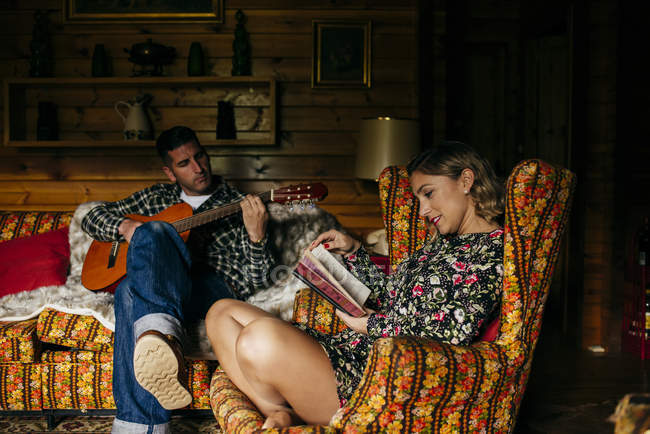 Frau sitzt im Sessel und liest Buch, während Mann zu Hause auf dem Sofa Gitarre spielt — Stockfoto