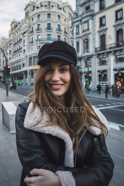 Femme brune souriante au chapeau élégant regardant la caméra sur la scène de la rue — Photo de stock