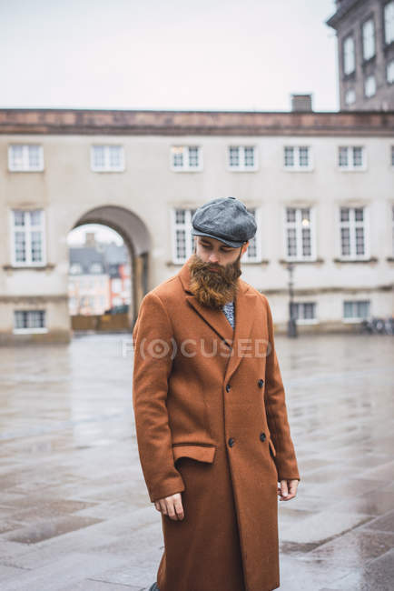 Стильный бородатый мужчина позирует на городской площади и смотрит вниз — стоковое фото