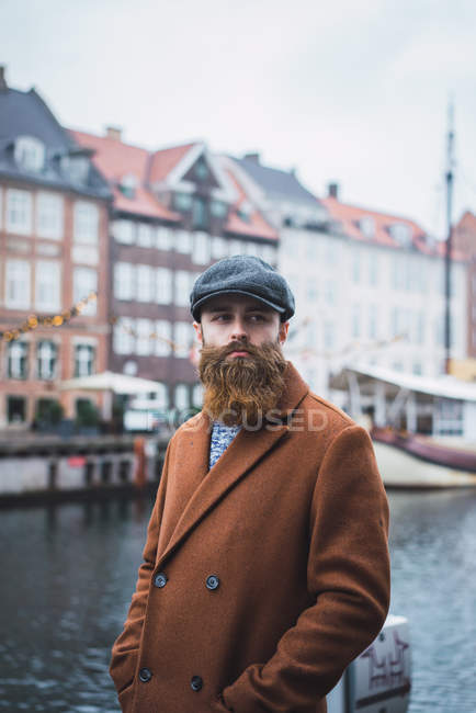 Front view of bearded man in coat and cap standing at riverside on city street and looking away — Stock Photo
