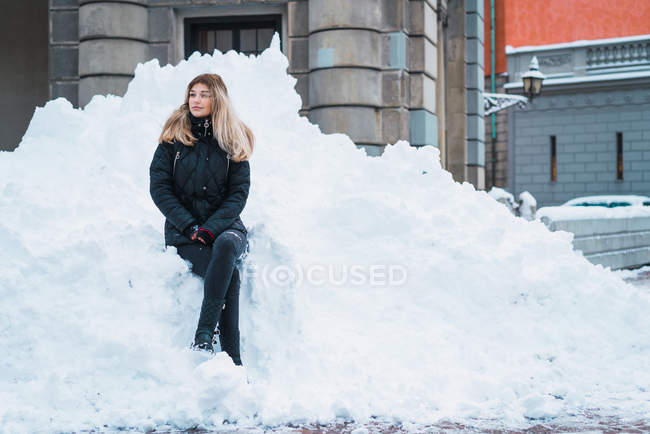 Молода красива жінка в повсякденному теплому одязі, сидячи на сніговій купі на вулиці і дивлячись — стокове фото