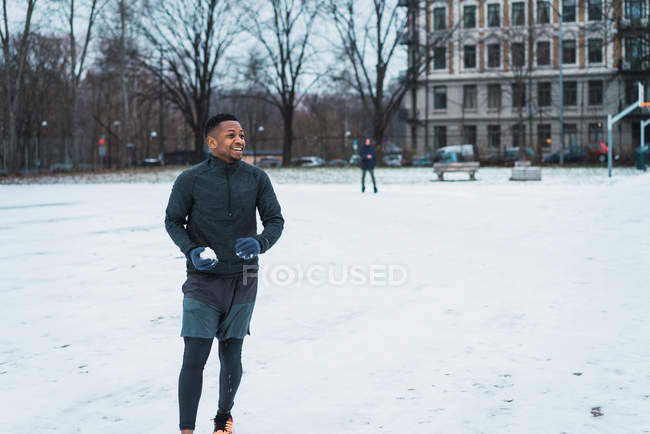 Вид спереду усміхненого спортсмена в теплому одязі, що стоїть зі сніжкою на дитячому майданчику . — стокове фото