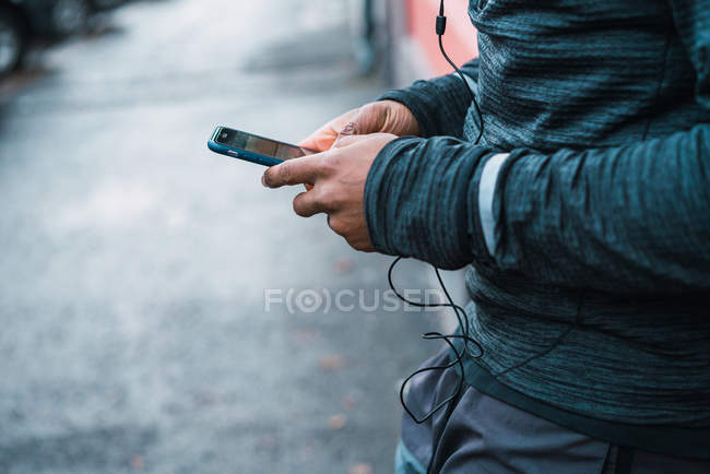 Mãos masculinas de colheita usando smartphone com fones de ouvido — Fotografia de Stock