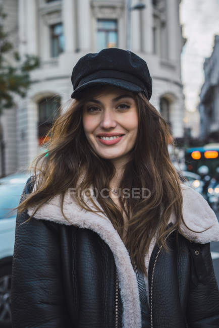 Femme souriante en chapeau élégant regardant la caméra sur la scène de la rue — Photo de stock