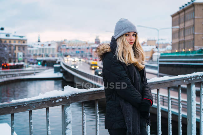 Vista lateral de la mujer joven en ropa de abrigo de pie en el puente en la ciudad de invierno y mirando a la cámara - foto de stock