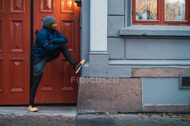 Homem esportivo inclinado com perna na parede enquanto aquece e alonga os músculos — Fotografia de Stock