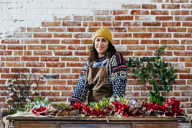 Retrato de mujer sonriente en sombrero y suéter de pie cerca de la mesa en atelier floral y mirando a la cámara . - foto de stock
