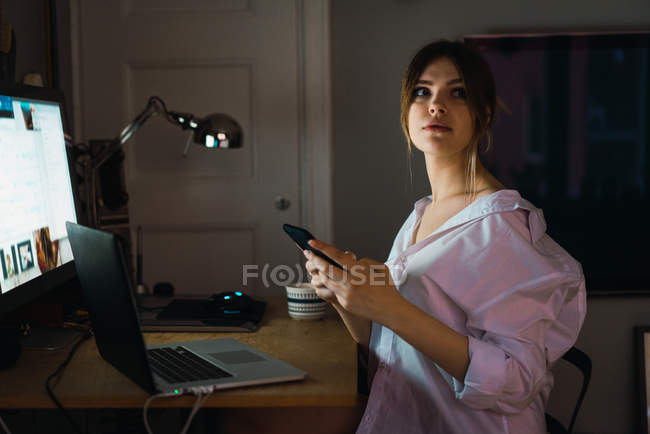 Woman sitting at laptop with smartphone in hands and looking away — Stock Photo