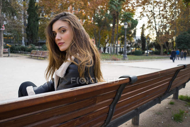 Retrato de mujer morena sentada en el banco en el parque y mirando por encima del hombro a la cámara - foto de stock