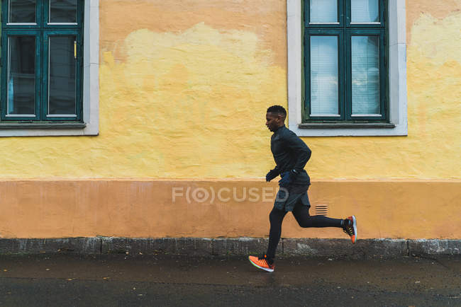 Боковой вид человека в теплой спортивной одежде, бегущего вдоль фасада здания — стоковое фото