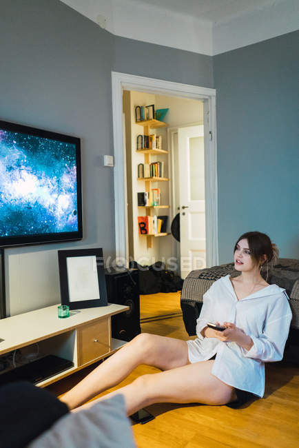 Woman sitting with smartphone on floor and looking at tv display on wall at home — Stock Photo