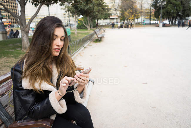 Молода красива жінка сидить на лавці і використовує смартфон у міському парку . — стокове фото