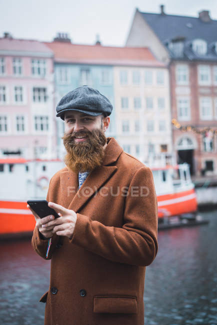 Retrato del hombre sonriente navegando por el teléfono inteligente en el río en la ciudad y mirando a la cámara - foto de stock