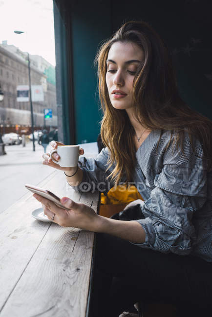 Retrato de chica morena bebiendo café en la cafetería y navegando por el teléfono inteligente - foto de stock