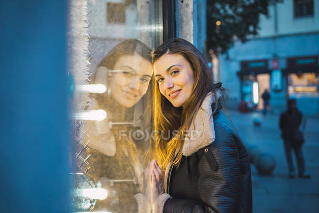 Side view of pretty woman looking at camera and leaning on glass of shop case on city street. — Stock Photo