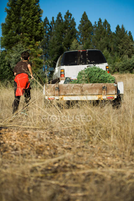 Back view of boy running to car with trailer and fir trees. — Stock Photo