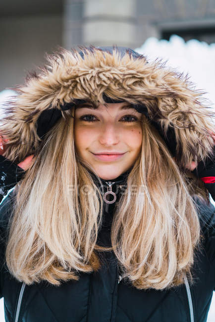 Portrait de femme blonde souriante à capuche avec des fourrures femme posant en ville d'hiver et regardant la caméra — Photo de stock