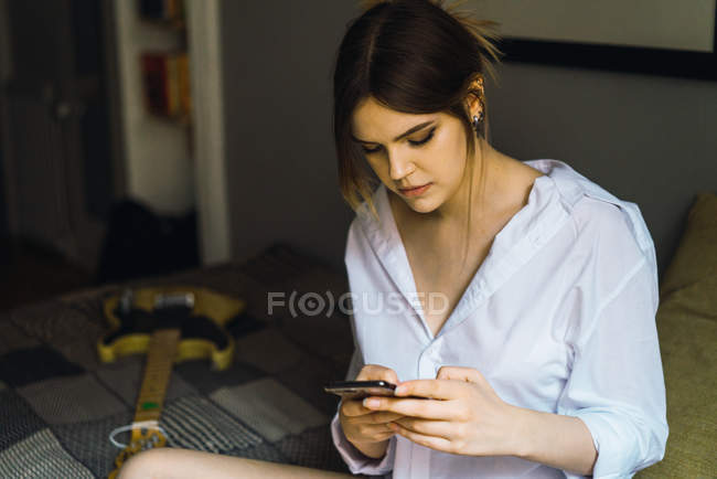 Mulher sentada na cama ao lado de guitarra e smartphone de navegação — Fotografia de Stock