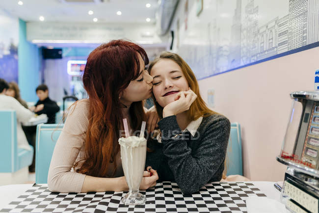 Portrait of redhead girl kissing girlfriends cheek at cafe table with milk shake — Stock Photo