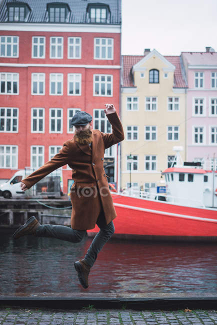 Portrait of bearded man in coat and cap gesturing in jump at riverside in city. — Stock Photo