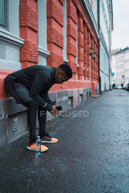 Jogador esportivo inclinado na fachada com o smartphone na mão descansando depois de correr — Fotografia de Stock