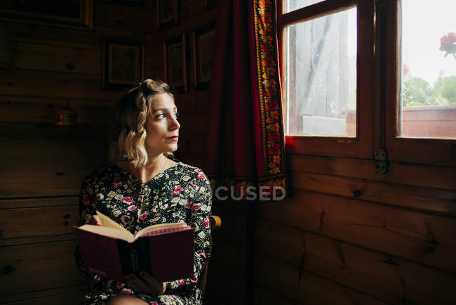 Retrato de mulher sonhadora olhando para a janela enquanto lê livro em casa . — Fotografia de Stock
