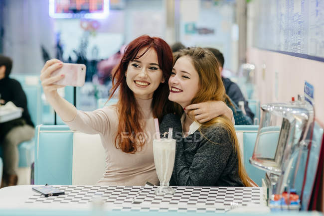 Retrato de chicas sonrientes haciendo selfie mientras están sentadas en la cafetería - foto de stock