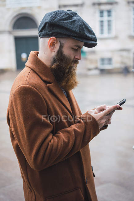 Side view of bearded man in coat and cap using phone on street — Stock Photo