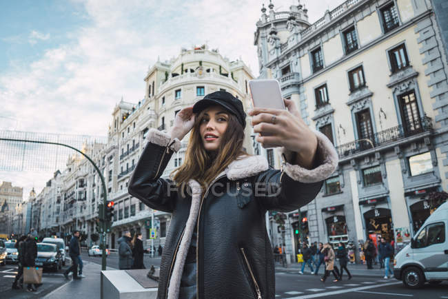 High angle view of brunette woman in cap taking selfie on smartphone at street scene — Stock Photo
