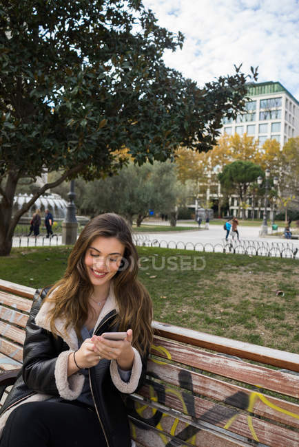 Portrait de femme brune souriante avec smartphone sur banc de parc — Photo de stock