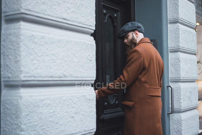 Vista lateral del hombre barbudo reflexivo en abrigo y tapa que abre la puerta de entrada - foto de stock