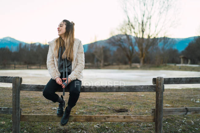 Retrato de una joven sentada en una cerca rural en el campo y mirando hacia otro lado - foto de stock